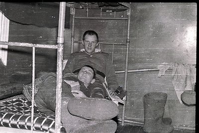 Two individuals seated on bunk beds in a confined, utilitarian space, likely a barracks or dormitory. The man on top holds a ...