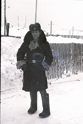 Winter uniform with fur collar and belt, holding mittens. Snow-covered fence and ground. Mid-20th century Soviet-era attire.