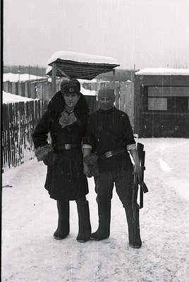 Two uniformed soldiers pose in winter attire, likely WWII-era German Wehrmacht. Snow-covered ground and barbed wire fence sug...