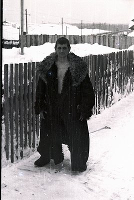 Portrait of a man in a heavy fur-lined coat, standing in snow-covered urban setting. Wooden fence and residential buildings i...
