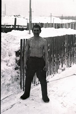 Young boy in mid-20th-century winter attire—shirtless with long wool socks and heavy boots—poses beside a snow-laden wooden f...