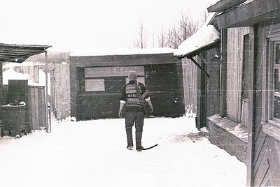 A man in a winter uniform, likely a postal worker, stands in a snowy alleyway holding mail. The scene features rustic wooden ...
