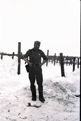 Soldier in winter gear poses by barbed-wire fence in snowy terrain, mid-20th century. Uniform suggests military training or g...