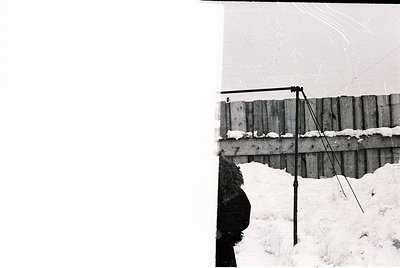 Black-and-white photograph of a rustic wooden fence with a slanted roof structure, likely a barn or storage shed. Snow covers...