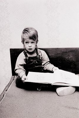 Young child in mid-20th-century overalls and slippers engrossed in an open book on a carpeted floor, seated beside a dark woo...