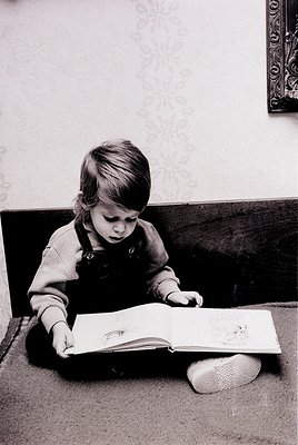 A young child engrossed in an open book on a dark carpeted floor, dressed in a long-sleeved shirt with suspenders and white s...