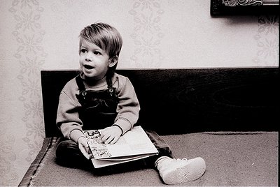 Black-and-white portrait of a young child (approx. 3-5 years) engrossed in a book, seated on a wooden bench. Overalls with bu...