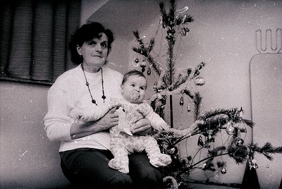 A woman cradles a baby beside a modestly decorated Christmas tree in an indoor setting, likely mid-20th century. The tree fea...