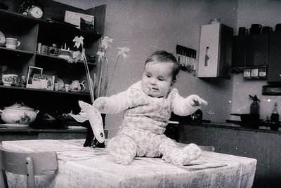 Vintage black-and-white photo of a toddler in a floral onesie, seated on a wooden stool, holding a paper airplane. Surroundin...