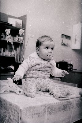 Vintage black-and-white photo of a toddler sitting on a patterned tablecloth, holding a toy. Indoor setting with floral wall ...