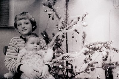 A mid-20th century (likely 1960s–1970s) black-and-white holiday scene: an older child holds a baby in front of a modestly dec...