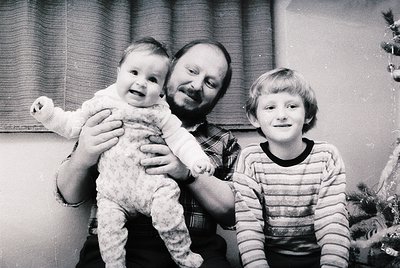 Family portrait from the 1970s–1980s, featuring a man holding a toddler while a young boy stands beside him. Indoor setting w...