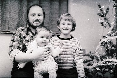 Family portrait from the 1970s–1980s featuring a man in a plaid shirt holding a baby, with a smiling boy in striped sweater b...