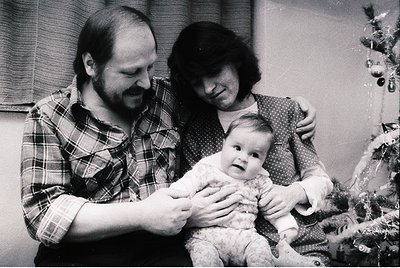 Family portrait featuring a man in a plaid shirt, woman in a patterned blouse, and a baby in a knitted outfit, posed indoors ...