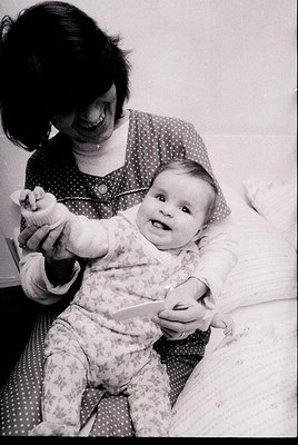 A tender black-and-white moment: a woman in a polka-dotted dress cradles an infant in floral pajamas, both smiling. Mid-20th ...