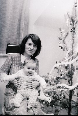 A woman in a patterned dress holds a baby in a festive indoor setting, likely during the 1970s. A decorated Christmas tree wi...