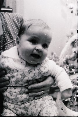 Black-and-white portrait of an infant in mid-1960s attire, wearing a patterned long-sleeve bodysuit and knitted sweater. The ...