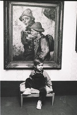 Vintage portrait of a child seated on a simple wooden stool, posing beside a framed painting of a woman in period attire (195...
