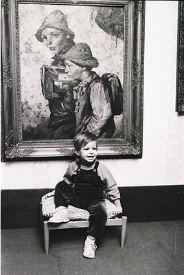 Vintage portrait of a young boy seated on a stool before a framed painting of a woman in outdoor attire, likely 1950s–1960s. ...