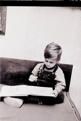 Young child engrossed in a book on a wooden bench, mid-20th century. Overalls, slippers, and a classic hardcover suggest a no...
