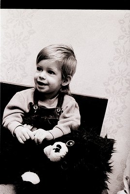 Young child in 1970s-style overalls holding a vintage teddy bear with fur trim, seated indoors against patterned wallpaper. C...