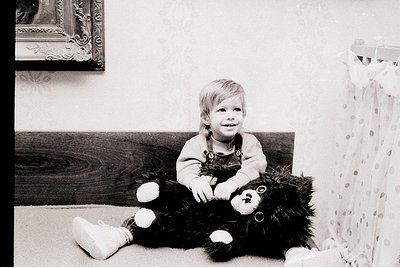 Vintage black-and-white portrait of a young child hugging a large teddy bear in a cozy indoor setting. Wooden headboard and f...
