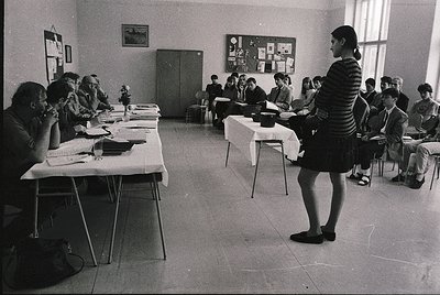 Classroom setting with students seated at long tables, engaged in a lecture or discussion. A teacher in a striped dress stand...