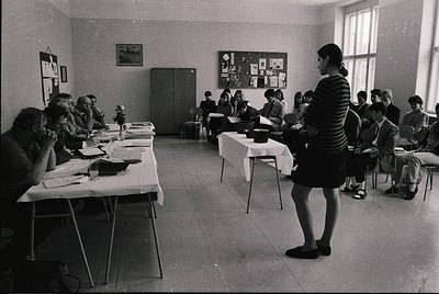 Classroom setting with mid-20th century institutional furniture, likely 1960s–1970s. Teacher in striped dress addresses seate...