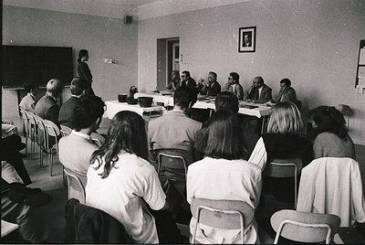 Formal conference or meeting in a mid-century institutional setting, likely 1960s–1970s. Group of seated attendees facing a p...