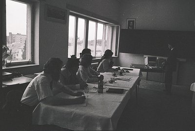 Vintage black-and-white classroom scene with five seated students focused on individual workstations. Long table with typewri...