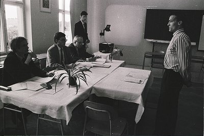 Mid-20th century conference room with four men in formal attire (1960s-1970s). White tablecloths, vintage projector, and clas...
