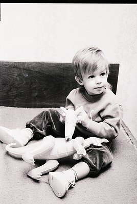 Vintage black-and-white photo of a young child (approx. 3-5 years) sitting on a carpeted floor, holding a stuffed duck toy. T...