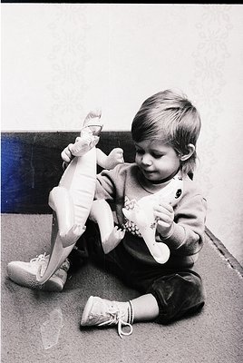 Young child in 1970s-style overalls and lace-up shoes plays with a white stuffed toy, seated on a textured carpeted floor. In...
