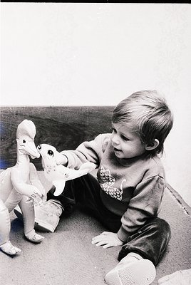 A young child in 1960s-era clothing plays with two vintage toy dinosaurs on a concrete surface. The child’s sweater features ...