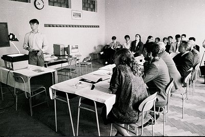 1970s classroom lecture hall with instructor at podium, students seated at long tables. Plain white walls, minimal decor, and...