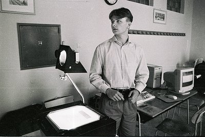 Man in a 1970s–80s office setting, standing beside a vintage slide projector and typewriter. White shirt, jeans, and a focuse...