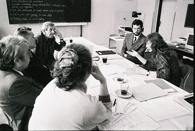 A 1970s-era office meeting featuring six professionals in formal attire around a rectangular table. Black-and-white chalkboar...