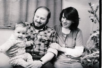 A black-and-white family portrait from the 1970s–1980s, featuring a man in a plaid shirt, a woman in a patterned blouse, and ...
