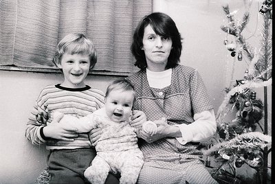 Family portrait featuring a woman holding a baby, with a young boy standing beside her. Indoor setting with a decorated Chris...