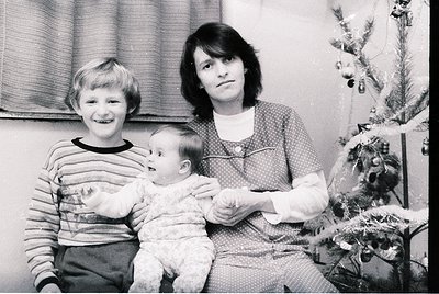 A 1970s family portrait featuring a woman holding a baby, with a smiling older child beside her. Indoor setting with a decora...