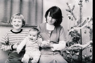 Family portrait featuring a woman holding a baby, with a young boy standing beside them. Decorated Christmas tree with orname...