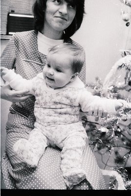 A woman in a polka-dot blouse holds a toddler in a floral onesie, both posing indoors near a decorated Christmas tree. The sc...