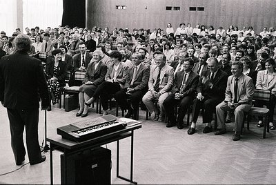 A formal indoor gathering in a large lecture hall or conference room, featuring a speaker addressing an attentive audience se...