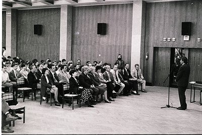 A formal indoor gathering in a mid-century conference hall, featuring a speaker at a podium addressing an attentive audience ...