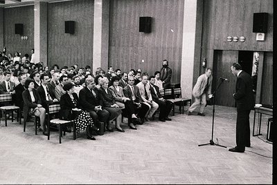 Mid-century lecture hall with tiered seating, featuring a speaker at a podium with microphone. Audience in formal attire, lik...