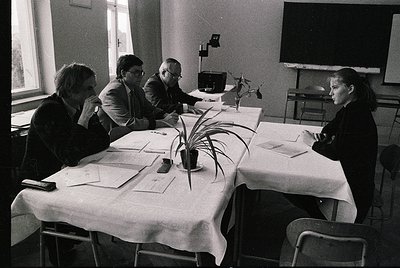 Four individuals seated around a rectangular table in a formal indoor setting, likely a conference or meeting room. The table...