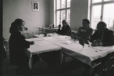 Four women in 1970s-era uniforms study architectural plans at a long table in a dimly lit classroom. Framed portrait above wi...
