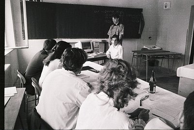 Classroom setting with students seated at desks, facing a teacher at a raised station. Blackboard with chalk writing, vintage...