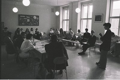 Mid-20th century classroom with U-shaped desks, students seated facing instructor. Blackboard displays solar system diagram. ...