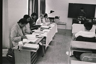 Black-and-white conference scene: six men seated at a rectangular table reviewing documents, surrounded by stacks of papers. ...